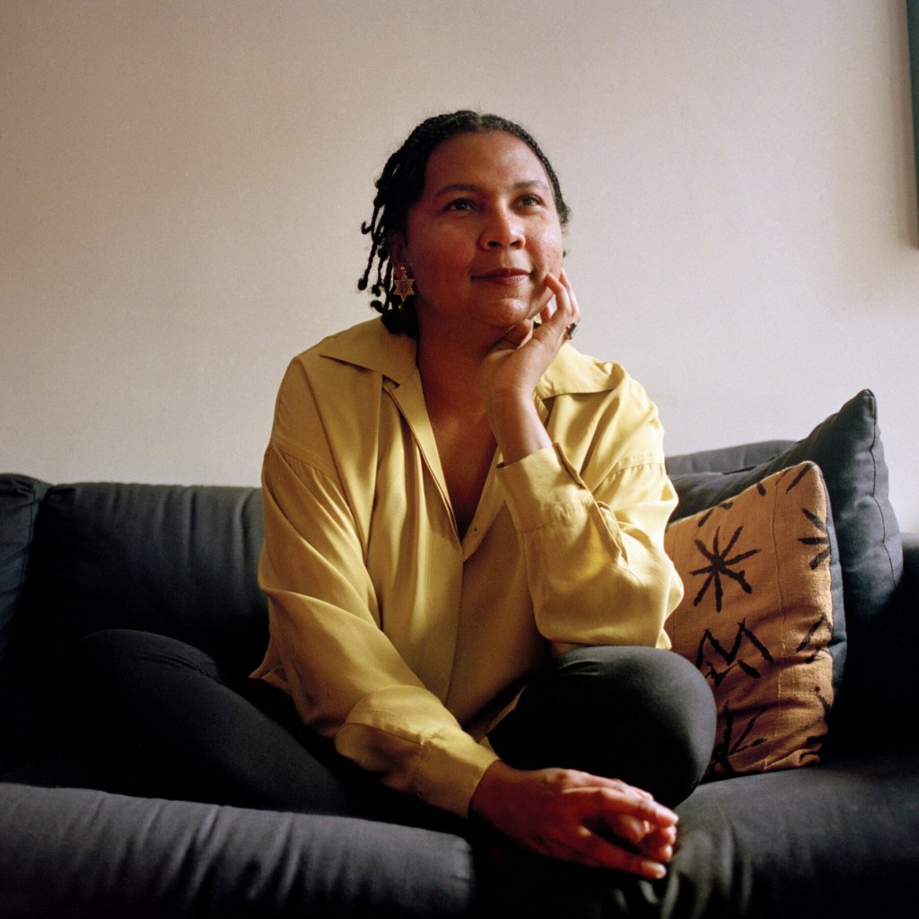 Image of author bell hooks with plaits in her hair, a gold blouse, and black pants, sitting on a sofa. 
